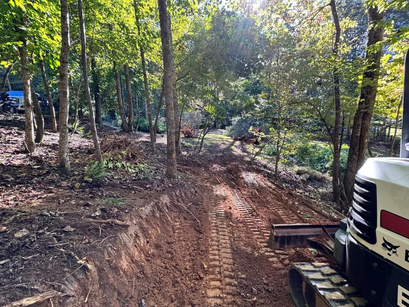 Freshly cut dirt access road through hardwoods with a mini excavator at the edge of the frame