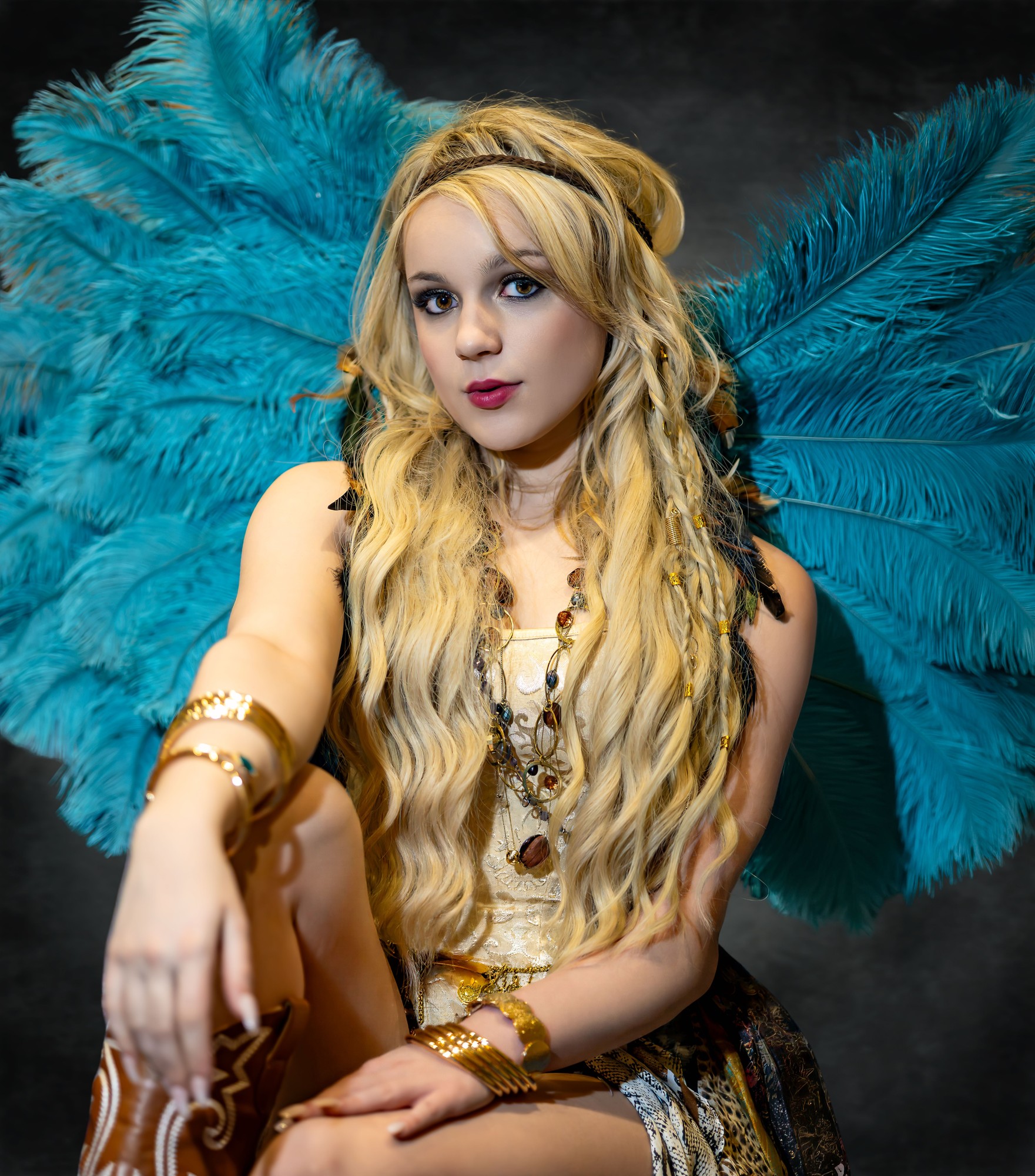 Model Sydney wearing tall teal ostrich-feather wings, a gold brocade top, layered gold jewelry and bangles, seated and looking directly into the camera