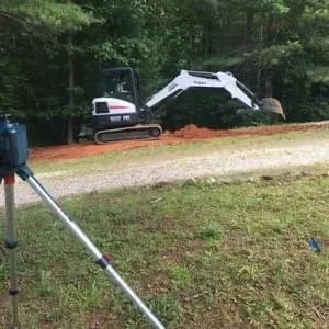 Mini excavator cutting into a hillside for a garage site with a hidden root cellar, with a survey level in the foreground